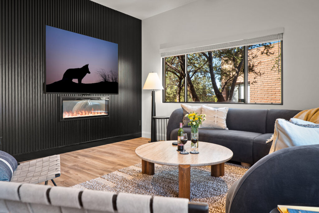Living room seating arranged around a linear fireplace set into a dark slat wall, with a window to the trees outside